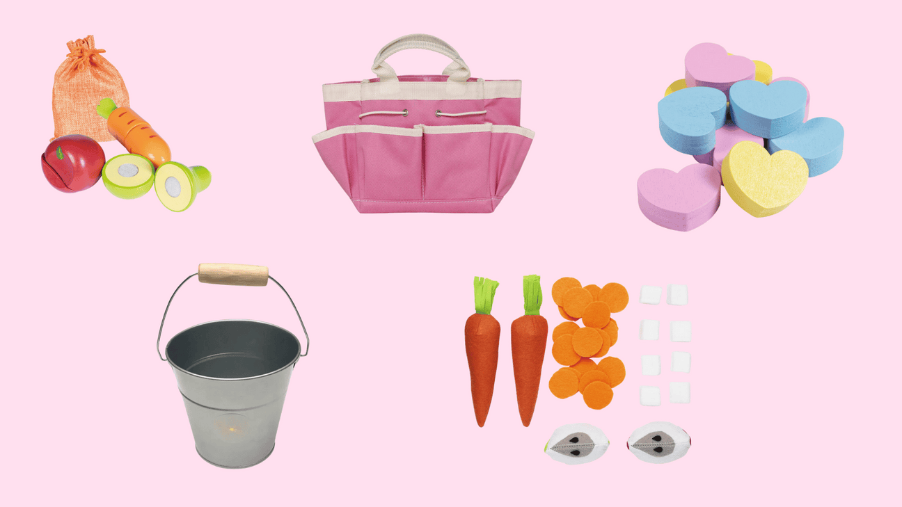 Verschiedene bunte Spielzeuge für Kinder auf rosa Hintergrund, darunter Holzgemüse, eine pinke Stofftasche, bunte Herzformen, ein Metalleimer und Filz-Zubehör.